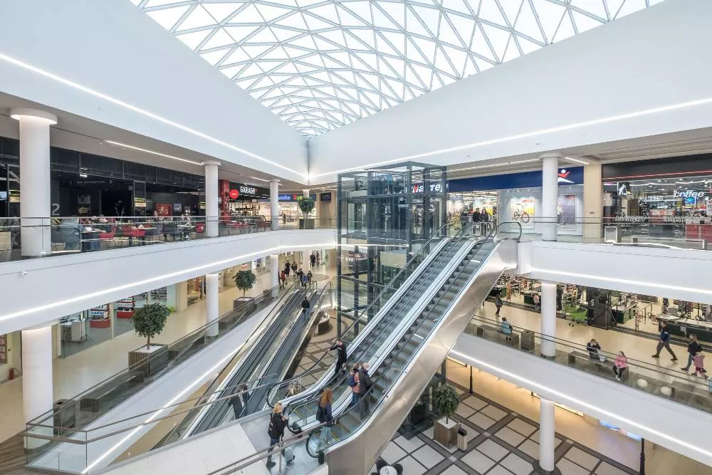 Intérieur lumineux d’un centre commercial moderne avec escalators, ascenseur vitré, boutiques et verrière en toiture.